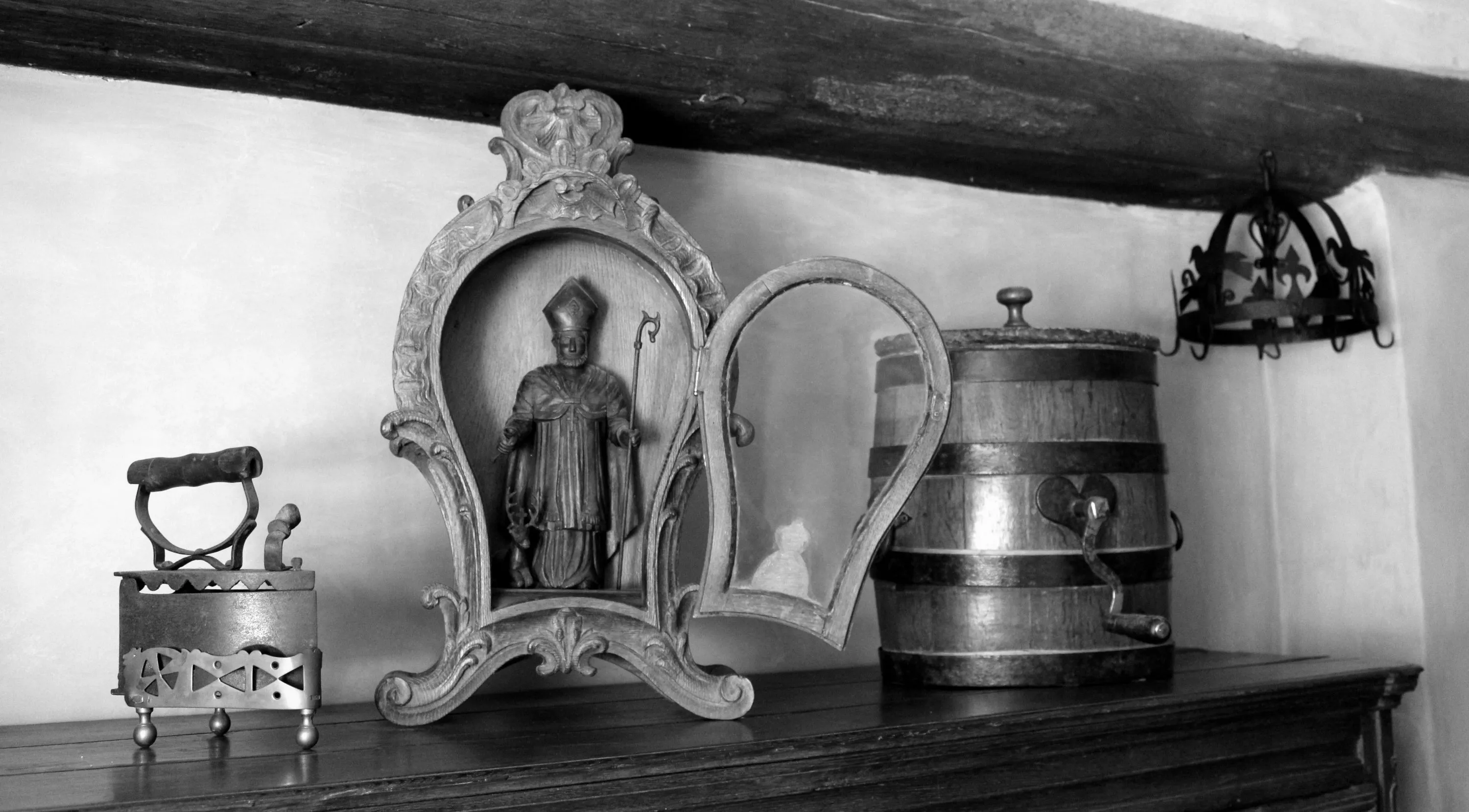 Sur un buffet du milieu du XIXe siècle se trouvent trois objets : un fer à repasser à braises en fonte posé sur son support en laiton ajouré, une niche vitrée en bois sculpté contenant une statue en bois de Saint Hubert et une baratte en chêne en forme de tonnelet décorée d’un cœur en fer. Une couronne d’office ornée alternativement de fleurs de lys et d’oiseaux est en arrière-plan. Ⓒ 2024 CC BY-NC 4.0 Gilles Pirlot de Corbion