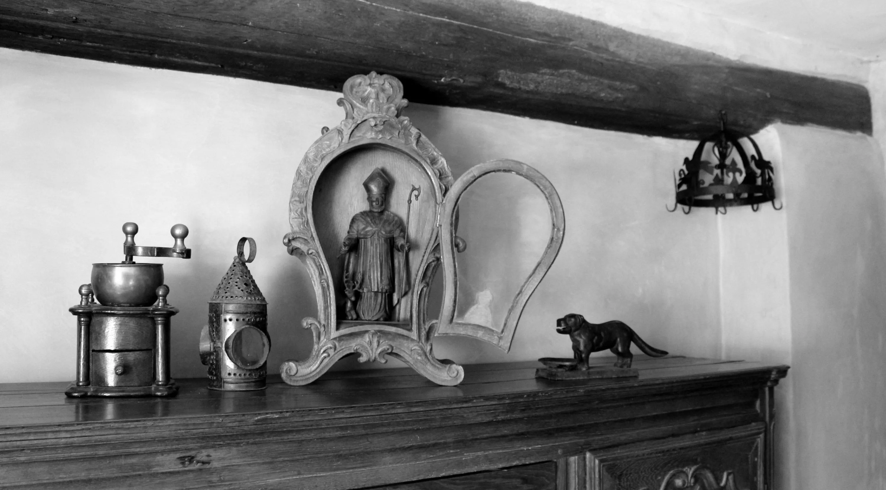 Quatre objets du XIXe siècle sont posés sur un buffet de cette époque : un moulin à café en chêne foncé à colonnettes, une lanterne portative à chandelle en tôle repoussée en fer étamé, une niche en chêne sculpté contenant une effigie de Saint Hubert, un casse-noix en fonte en forme de chien. En arrière-plan figure une couronne d’office du XVIIIe siècle. Ⓒ 2024 CC BY-NC 4.0 Gilles Pirlot de Corbion