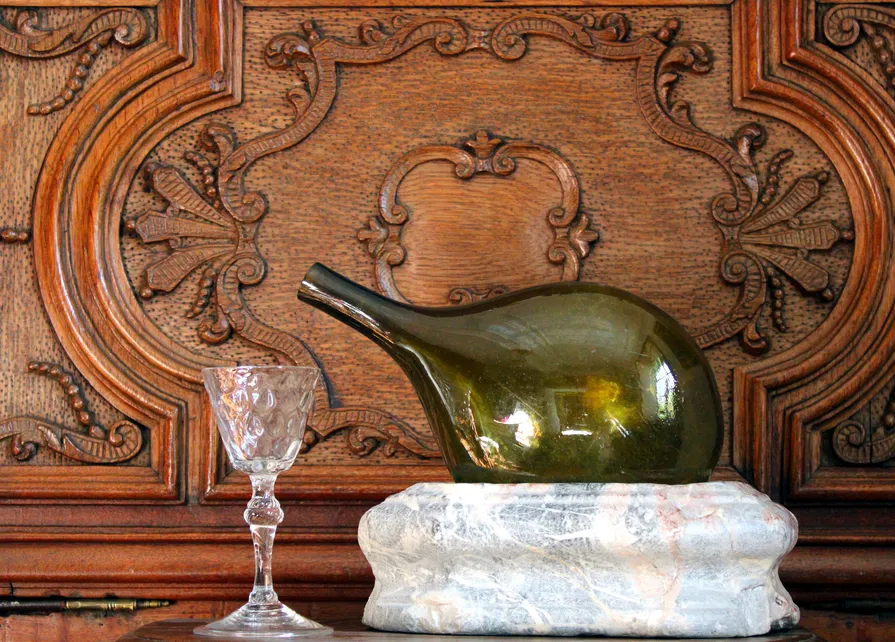 Une bouteille d’eau de Spa du XVIIIe siècle en verre soufflé vert olive posée sur un socle en marbre de Saint-Remy et un verre en cristal taillé du XVIIIe siècle d’origine liégeoise sont présentés devant un meuble en chêne sculpté aux motifs baroques créé par le maître-artisan Jean-Georges Scholtus durant la première moitié du XVIIIe siècle. Ⓒ 2025 CC BY-NC 4.0 Gilles Pirlot de Corbion