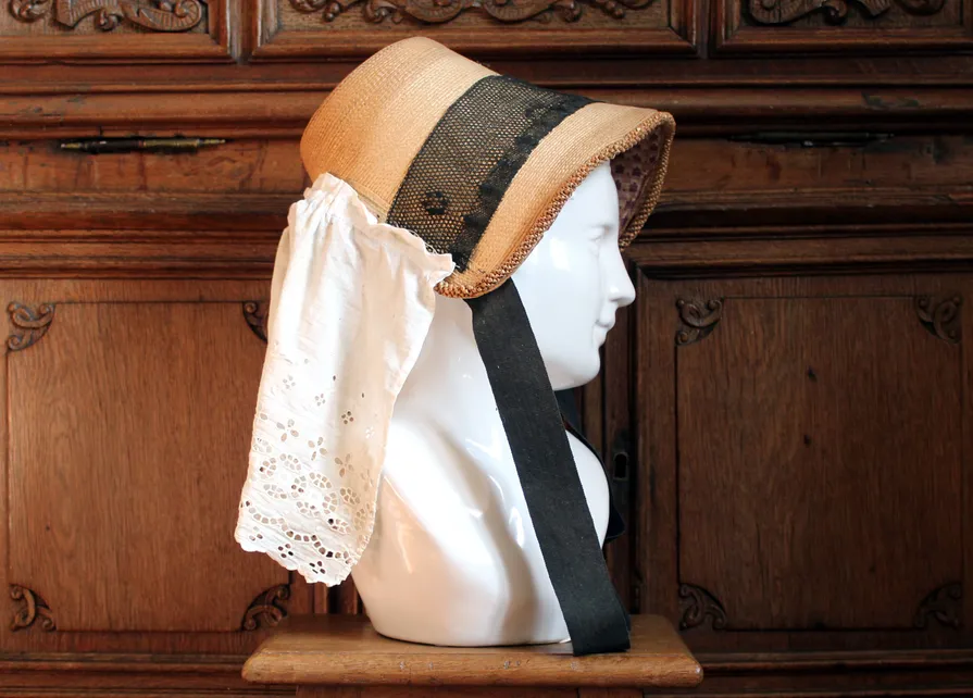Devant un meuble en chêne sculpté du XVIIIe siècle, un buste en porcelaine présente un barada (aussi appelé bavolet), chapeau traditionnel ardennais en paille, reconnaissable à son volant de tissu ou de dentelle protégeant la nuque et ayant donné son nom à l’ensemble. Devant un meuble en chêne sculpté aux motifs baroques créé par le maître-menuisier Jean-Georges Scholtus au début du XVIIIe siècle, buste en porcelaine blanche servant à présenter un barada, chapeau ardennais traditionnel, en paille, prolongé par un volant d'étoffe ou de dentelle pour protéger le cou du soleil. Ⓒ 2025 CC BY-NC 4.0 Gilles Pirlot de Corbion