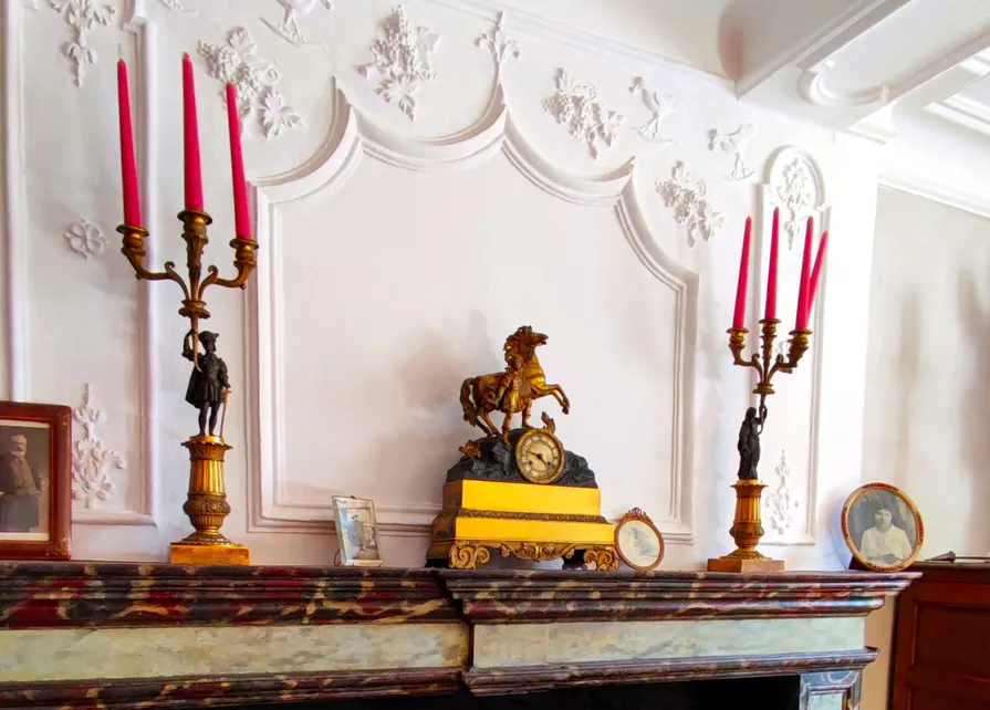 Photographies des aïeux de Gilles Pirlot de Corbion sur la cheminée de bois peinte en faux-marbre surmontée d’un trumeau stuqué dans la belle salle de la Blanche Maison le jour de son inauguration officielle. Le manteau de la cheminée, ayant conservé sa peinture faux-marbre d’origine, présente une pendule en bronze d’époque Charles X encadrée de ses deux candélabres assortis. Cette garniture de cheminée est entourée de photographies des aïeux de Gilles Pirlot de Corbion. À l’arrière-plan se dessine un rare trumeau en stuc où se mélangent des éléments de style Louis XIV et des motifs plus populaires tels que des pampres et des merles tenant une baie dans leur bec Ⓒ 2025 CC BY-NC 4.0 Didier Misson