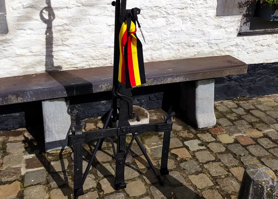 La crémaillère enrubannée aux couleurs de la Belgique attend d'être pendue dans la cuisine de la Blanche Maison avant son inauguration. Ⓒ 2025 CC BY-NC 4.0 Didier Misson Posée devant l’entrée de la Blanche Maison, la crémaillère du XVIIe siècle provenant de Regné est enrubannée aux couleurs de la Belgique