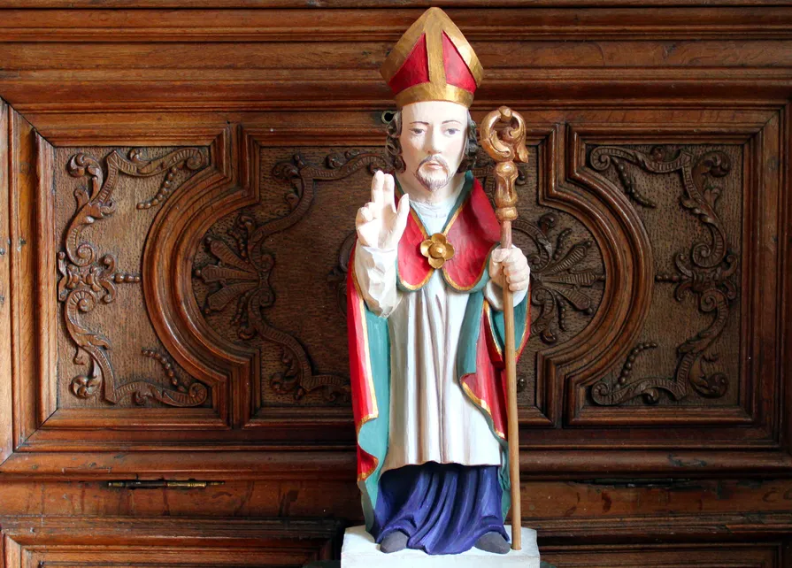 Statue de saint Hubert représenté en évêque bénissant, en chêne polychrome, copie agrandie d’une statue du XVIIIe siècle représentant saint Hubert de Liège, provenant de la chapelle de Harzy (Bastogne), conservée aujourd’hui au Musée de la Grande Ardenne. À l’arrière-plan, détail d’un meuble en chêne moyen à abattant du début du XVIIIe siècle attribué à l’atelier Scholtus de Bastogne. Ⓒ 2026 CC BY-NC 4.0 Gilles Pirlot de Corbion