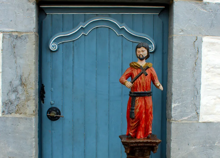 Posée sur une colonne décorée de torsades de fleurs et de fruits devant la porte d’entrée de la Blanche Maison à Nisramont, statue polychrome du XVIIIe siècle de facture naïve représentant saint François d'Assise debout, nu-tête et barbu portant un froc de moine rouge noué à la taille par une cordelière. Ⓒ 2026 CC BY-NC 4.0 Gilles Pirlot de Corbion