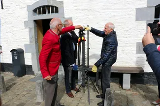 L'authentique crémaillère du XVIIe siècle, provenant de Regné (Vielsalm), est déplacée vers la cuisine sous l’œil attentif de Gilles Pirlot de Corbion. Ⓒ 2025 CC BY-NC 4.0 Didier Misson Jacques Wanlin et Emmanuel Rogman s’apprêtent à déplacer la crémaillère vers la cuisine