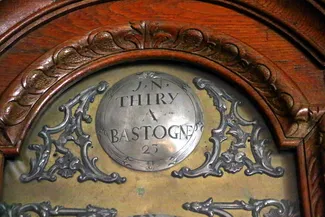 Plaque métallique ronde avec une inscription gravée en lettres capitales, J. N. THIRY A BASTOGNE, fixée sur un support en bois sculpté dans la partie supérieure de l’horloge ancienne présentée dans la belle salle de la Blanche Maison Ⓒ 2025 CC BY-NC 4.0 Didier Misson