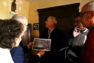 Après un décapage méticuleux, le bouquet en stuc avec sa console destinée à une statuette religieuse a retrouvé son éclat d’antan. Ⓒ 2025 CC BY-NC 4.0 Didier Misson Michel Francard tient une photographie de 2011 qui montre l’état dans lequel se trouvait le bouquet en stuc situé au-dessus de la porte de la chambre derrière le feu