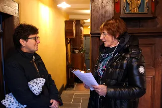 Conversation entre la représentante des sonneurs de Bertrix et Thérèse Colla, une habitante de Nisramont qui est la mémoire vivante du village, dans le corridor de la Blanche Maison lors du goûter de Noël le 20 décembre 2025.