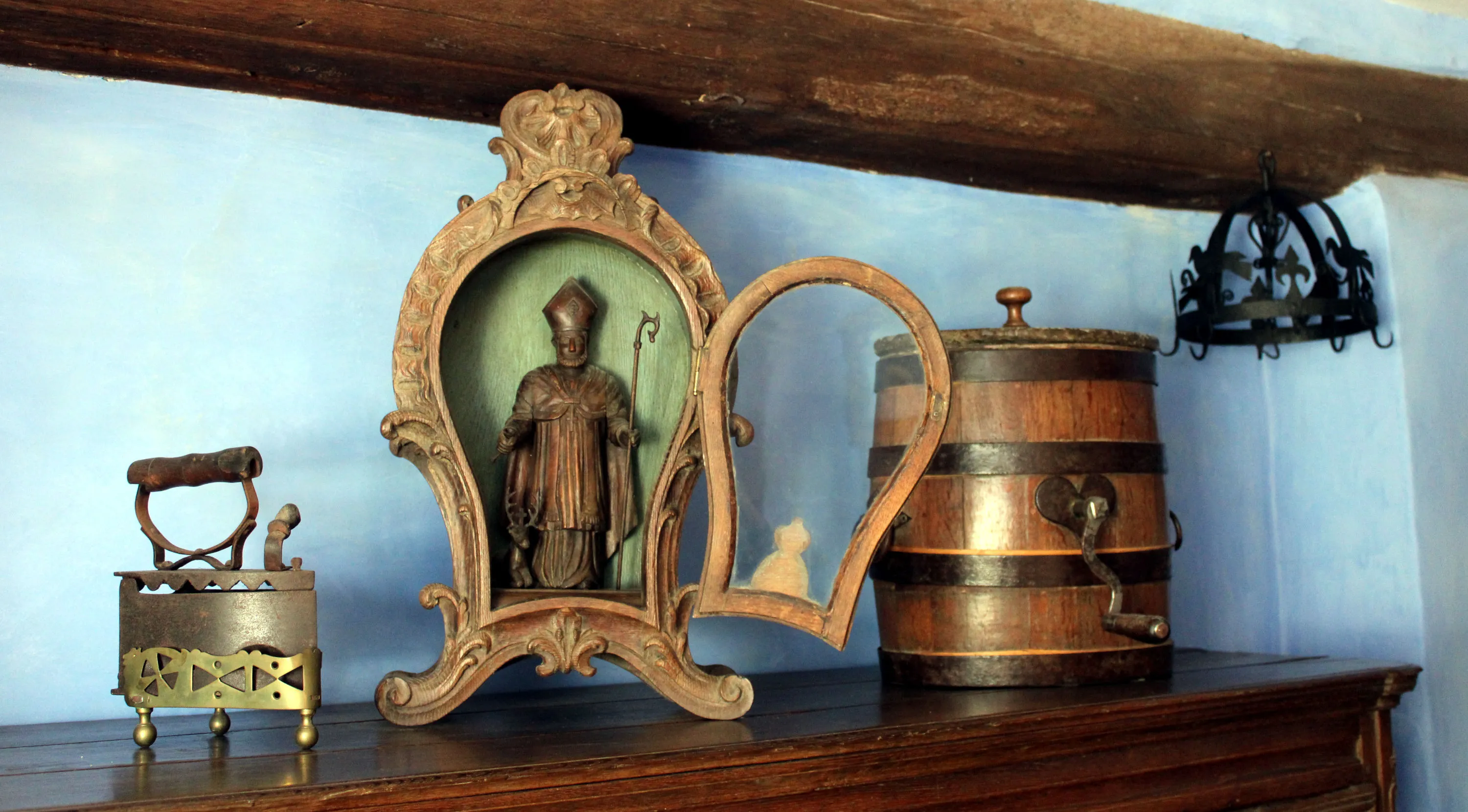 Sur un buffet du milieu du XIXe siècle se trouvent trois objets : un fer à repasser à braises en fonte posé sur son support en laiton ajouré, une niche vitrée en bois sculpté contenant une statue en bois de Saint Hubert et une baratte en chêne en forme de tonnelet décorée d’un cœur en fer. Une couronne d’office ornée alternativement de fleurs de lys et d’oiseaux est en arrière-plan. Ⓒ 2024 CC BY-NC 4.0 Gilles Pirlot de Corbion