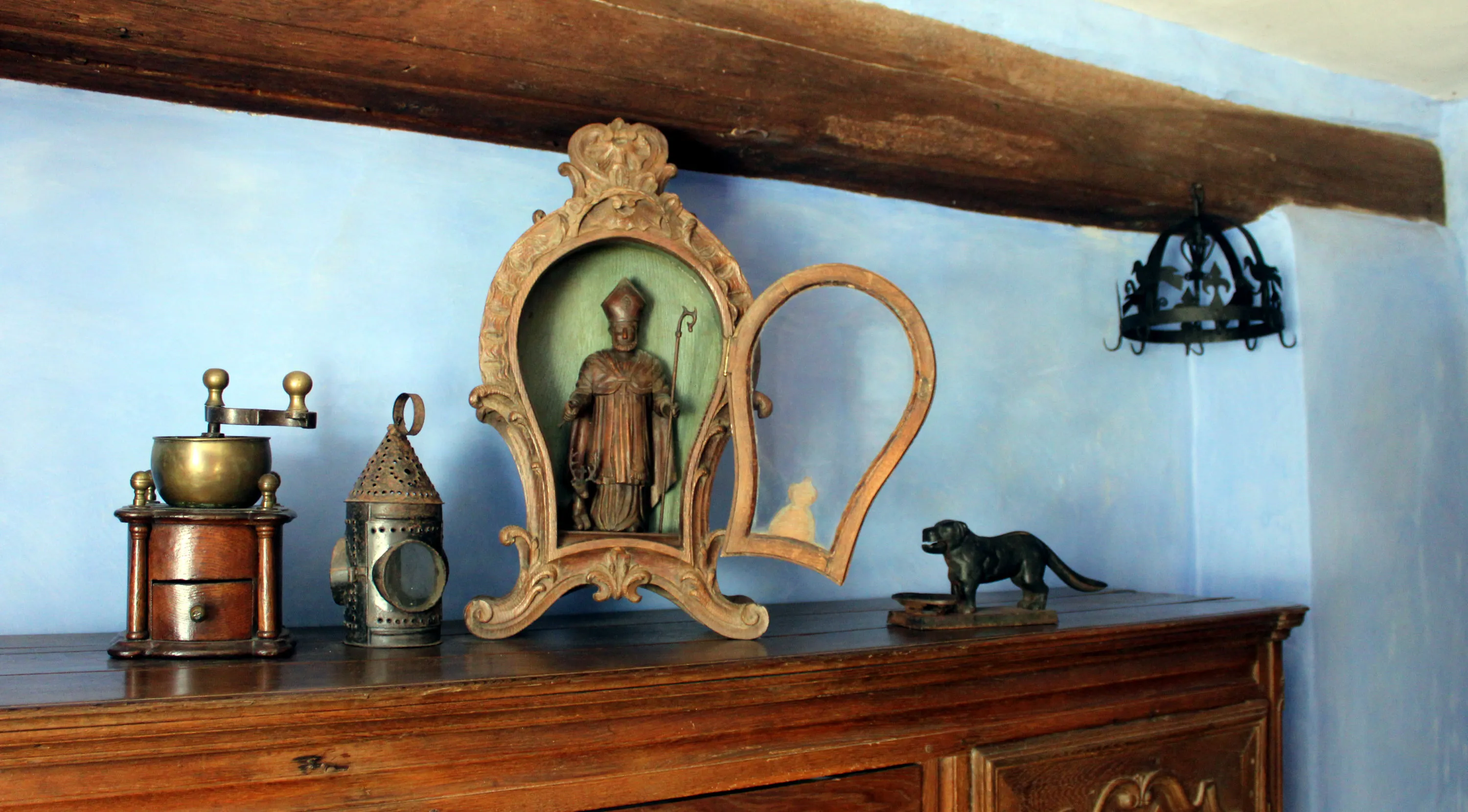 Quatre objets du XIXe siècle sont posés sur un buffet de cette époque : un moulin à café en chêne foncé à colonnettes, une lanterne portative à chandelle en tôle repoussée en fer étamé, une niche en chêne sculpté contenant une effigie de Saint Hubert, un casse-noix en fonte en forme de chien. En arrière-plan figure une couronne d’office du XVIIIe siècle. Ⓒ 2024 CC BY-NC 4.0 Gilles Pirlot de Corbion