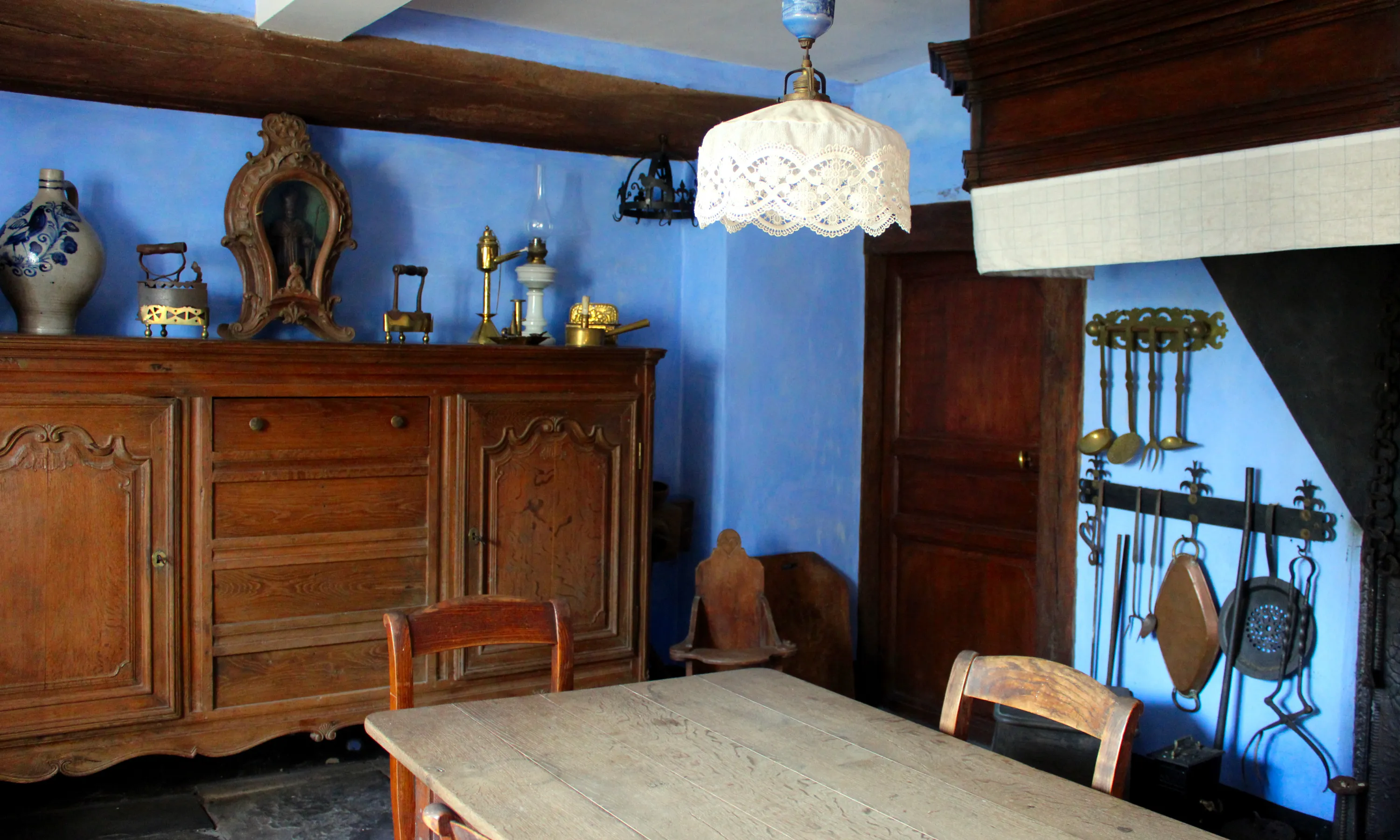 Le bleu très lumineux des murs de la cuisine résulte de l’ajout d’un pigment bleu outremer à la chaux dont ils sont badigeonnés. Le grand buffet provient de Mousny, un village voisin, ce qui en fait un meuble véritablement local. La table abrite un pétrin sous son plateau avec encore des traces de farine. De nombreux accessoires évoquent la vie quotidienne de jadis. Ⓒ 2024 CC BY-NC 4.0 Gilles Pirlot de Corbion