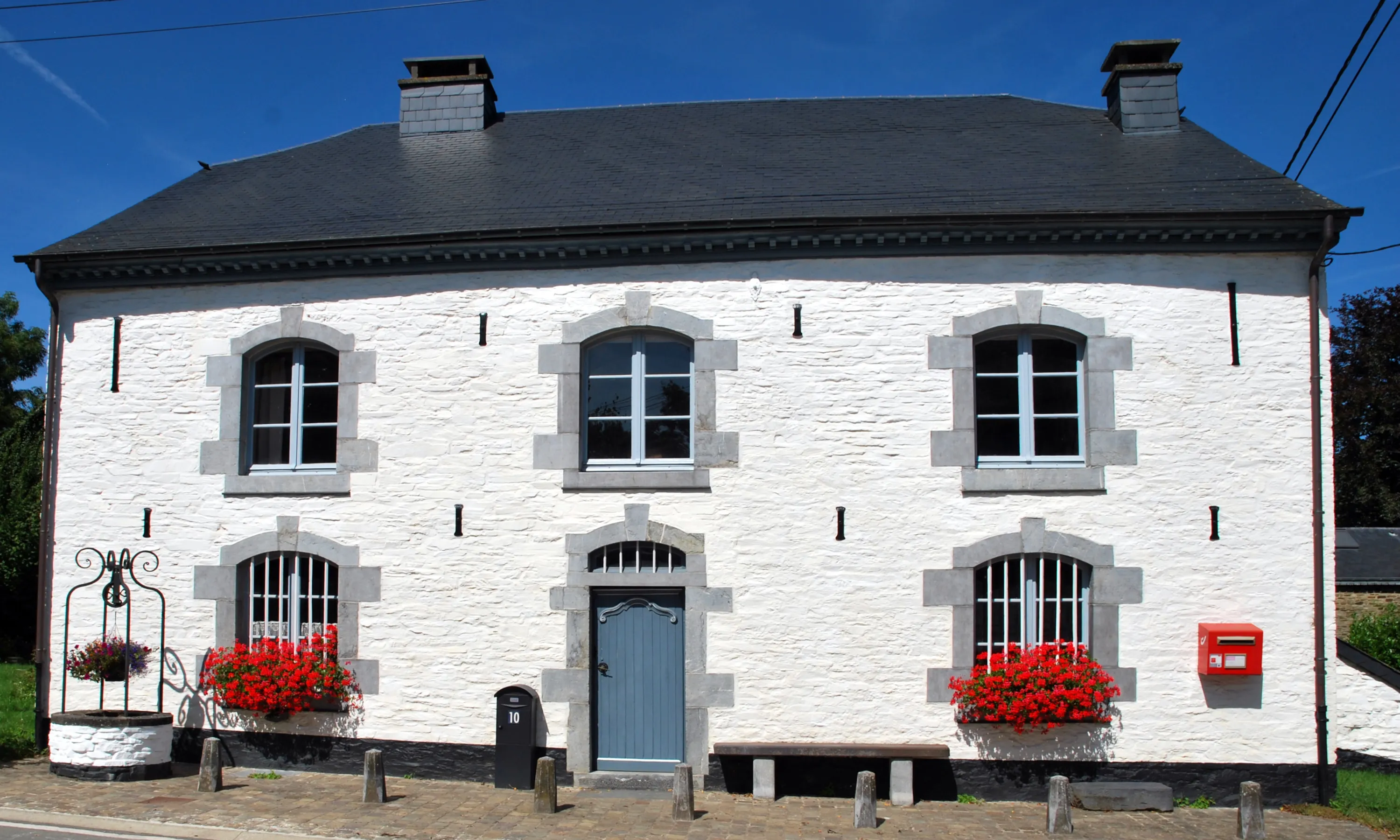 Façade en pierres chaulées de La Blanche Maison, un musée situé au centre de Nisramont en Ardenne belge. Cette façade comporte, au rez-de-chaussée, une porte d’entrée encadrée symétriquement de deux fenêtres munies de barreaux. A l’étage, trois fenêtres surmontent exactement les baies du rez-de-chaussée. Ⓒ 2024 CC BY-NC 4.0 Gilles Pirlot de Corbion