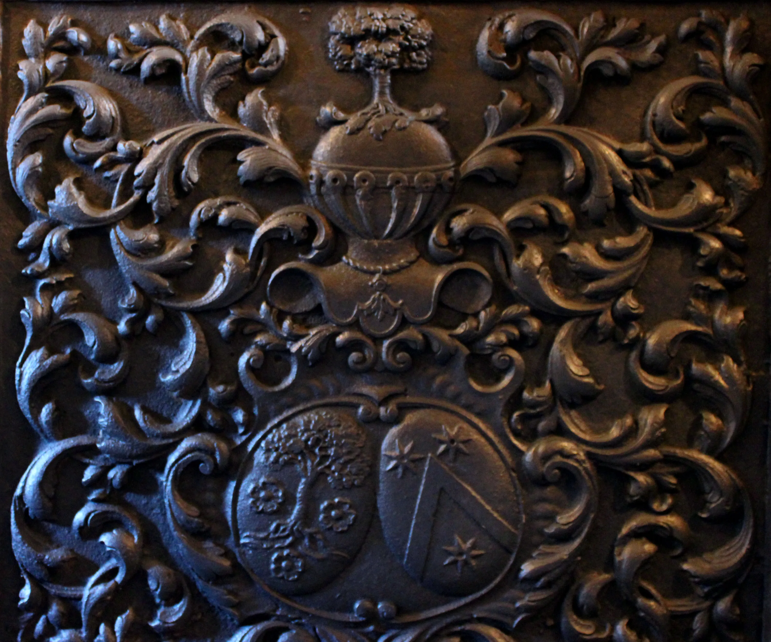 Cette taque en fonte est disposée en bas de l'escalier de la Blanche Maison. Un autre exemplaire de cette taque était exposé au Musée archéologique d'Arlon jusqu'au début des années 2000 mais il a été retiré des collections suite aux travaux de rénovation de l'institution muséale. Jeanne-Hyacinthe Poschet épousa Thomas Piret, seigneur de Saint-Ode et maître de forge, le 22 août 1685. Forgée dans la perspective de cette union, cette taque de cheminée à décor héraldique en fort relief arbore les armes d’alliance Piret de Sainte-Ode et Poschet surmontées d’un heaume avec cimier et de lambrequins. Ⓒ 2024 CC BY-NC 4.0 Gilles Pirlot de Corbion
