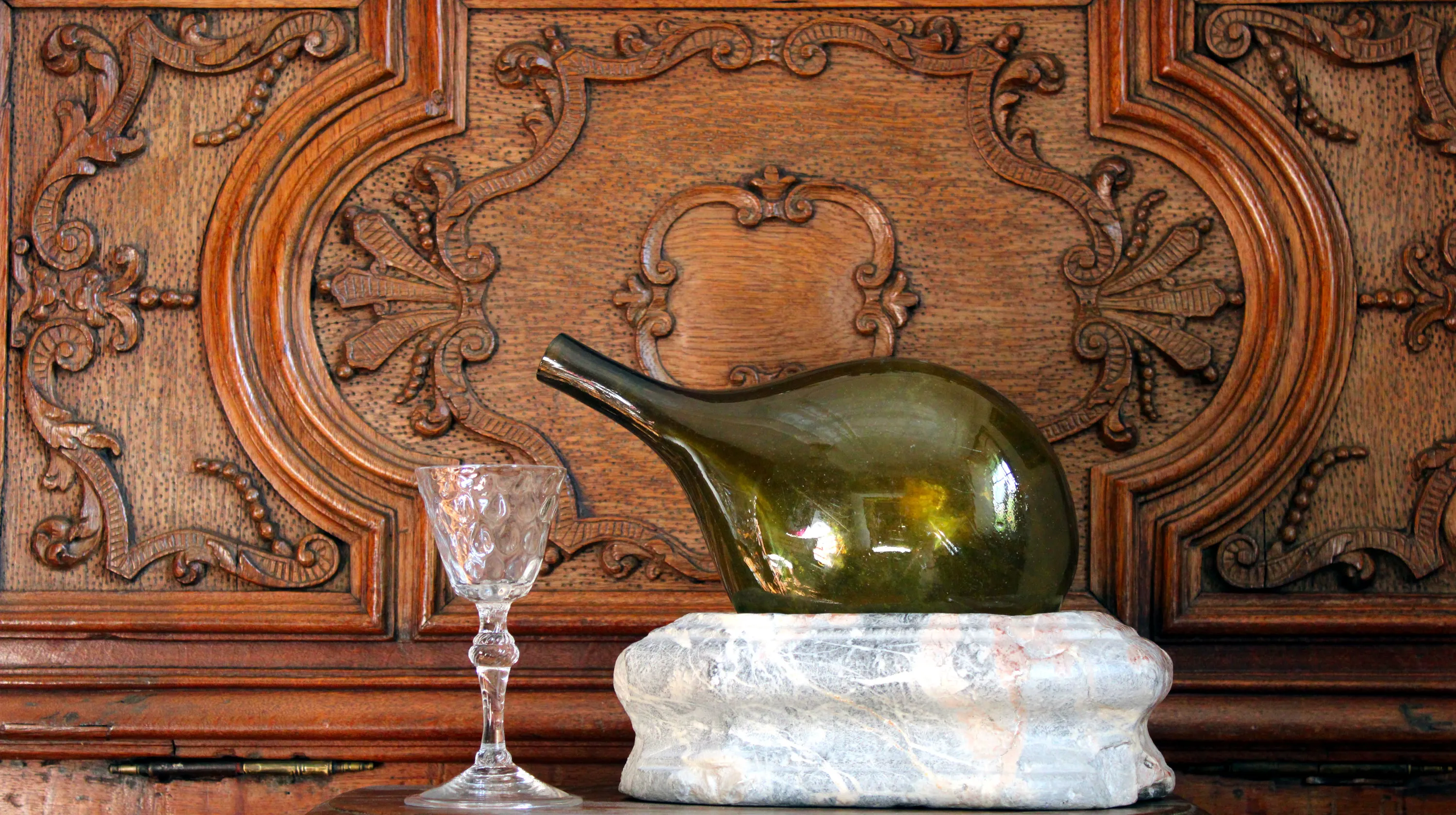 Une bouteille d’eau de Spa du XVIIIe siècle en verre soufflé vert olive posée sur un socle en marbre de Saint-Remy et un verre en cristal taillé du XVIIIe siècle d’origine liégeoise sont présentés devant un meuble en chêne sculpté aux motifs baroques créé par le maître-artisan Jean-Georges Scholtus durant la première moitié du XVIIIe siècle. Ⓒ 2025 CC BY-NC 4.0 Gilles Pirlot de Corbion