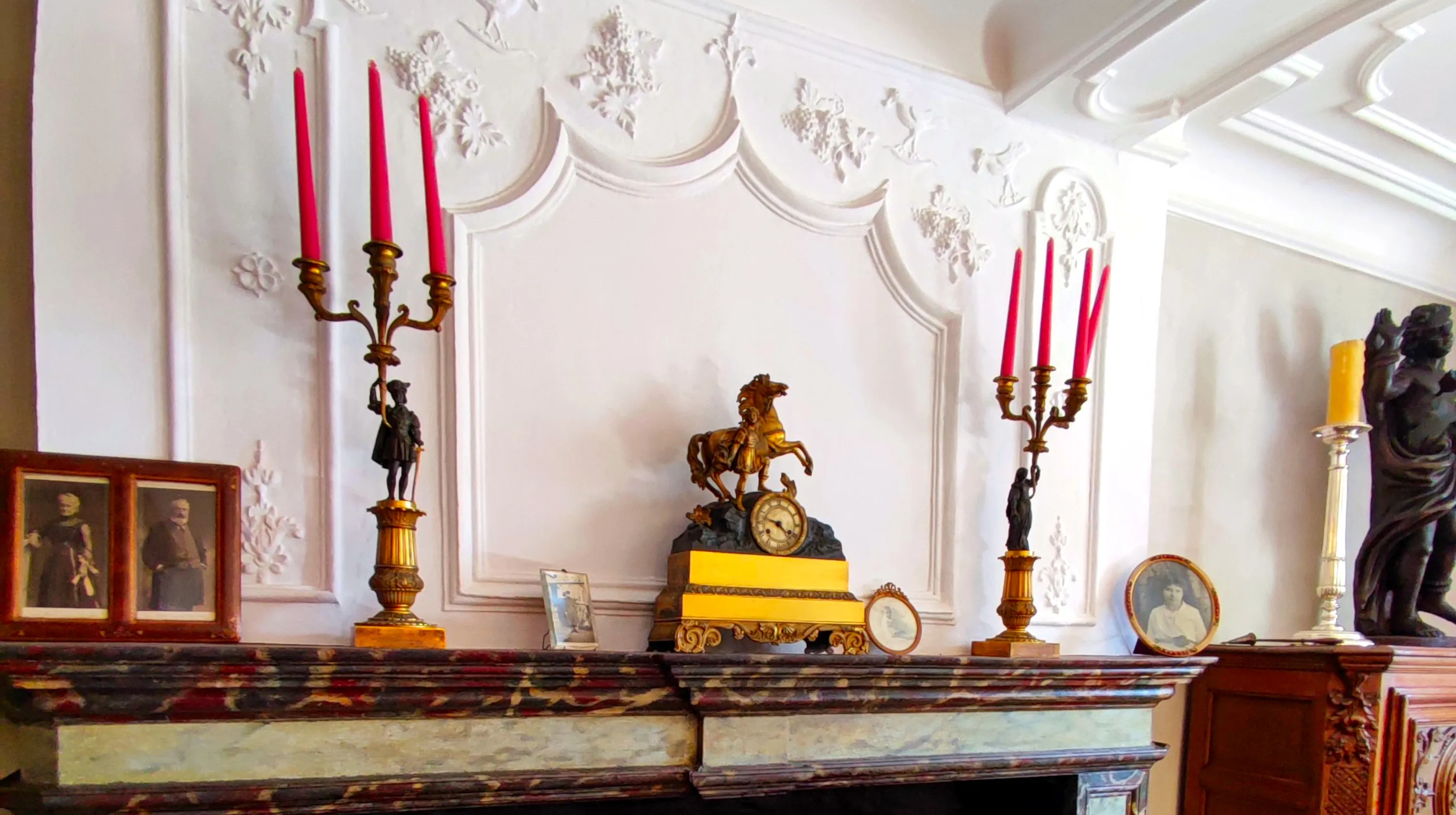 Le manteau de la cheminée, ayant conservé sa peinture faux-marbre d’origine, présente une pendule en bronze d’époque Charles X encadrée de ses deux candélabres assortis. Cette garniture de cheminée est entourée de photographies des aïeux de Gilles Pirlot de Corbion. À l’arrière-plan se dessine un rare trumeau en stuc où se mélangent des éléments de style Louis XIV et des motifs plus populaires tels que des pampres et des merles tenant une baie dans leur bec Ⓒ 2025 CC BY-NC 4.0 Didier Misson