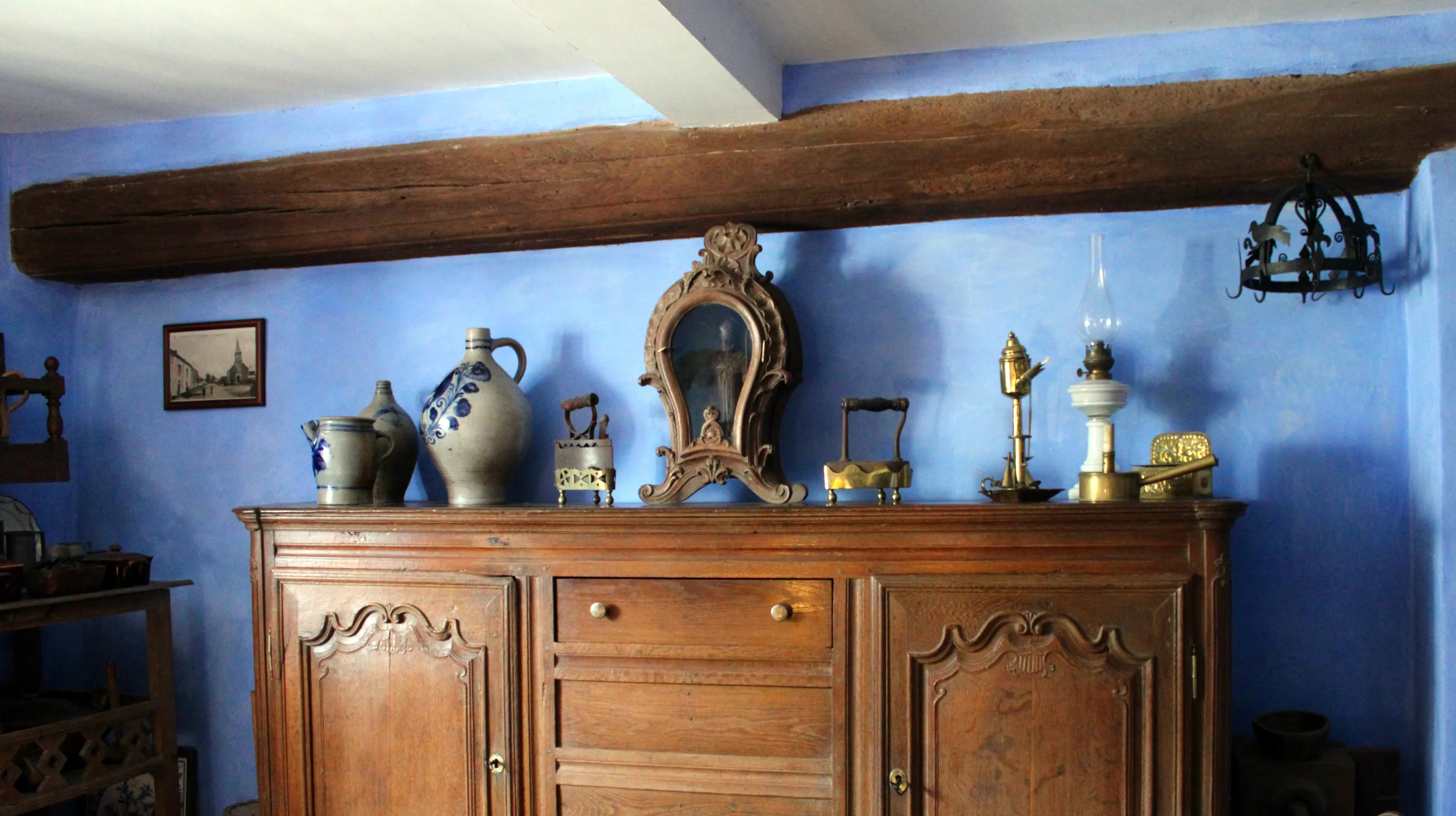 La cuisine chaulée en bleu outremer présente un buffet en chêne de couleur miel, provenant du village de Mousny, sur lequel sont posés des poteries en grès de La Roche-en-Ardenne, une niche vitrée de forme violonée contenant une statue de Saint Hubert, des fers à charbon et des luminaires anciens. Ⓒ 2026 CC BY-NC 4.0 Gilles Pirlot de Corbion