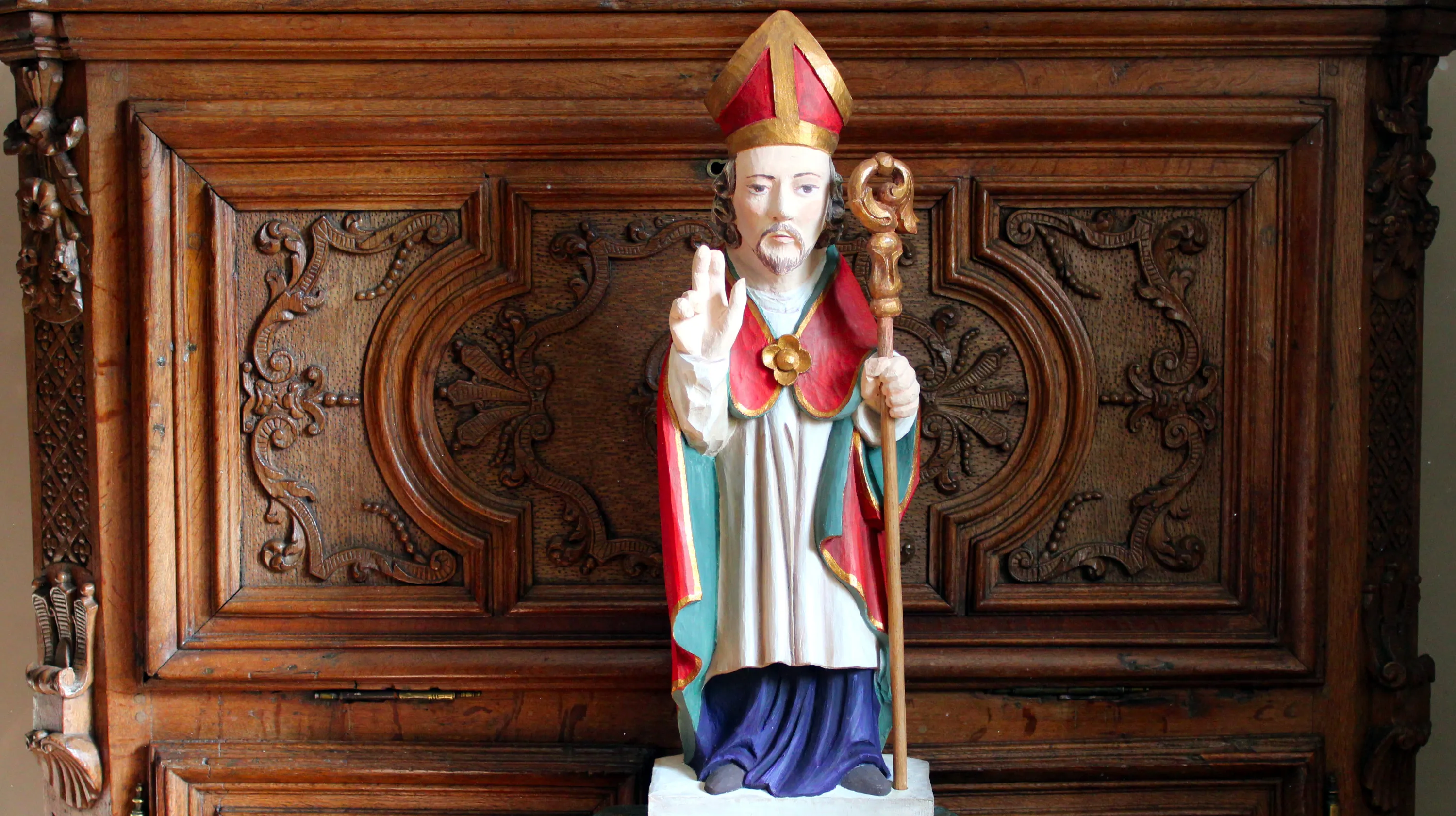 Statue de saint Hubert représenté en évêque bénissant, en chêne polychrome, copie agrandie d’une statue du XVIIIe siècle représentant saint Hubert de Liège, provenant de la chapelle de Harzy (Bastogne), conservée aujourd’hui au Musée de la Grande Ardenne. À l’arrière-plan, détail d’un meuble en chêne moyen à abattant du début du XVIIIe siècle attribué à l’atelier Scholtus de Bastogne. Ⓒ 2026 CC BY-NC 4.0 Gilles Pirlot de Corbion