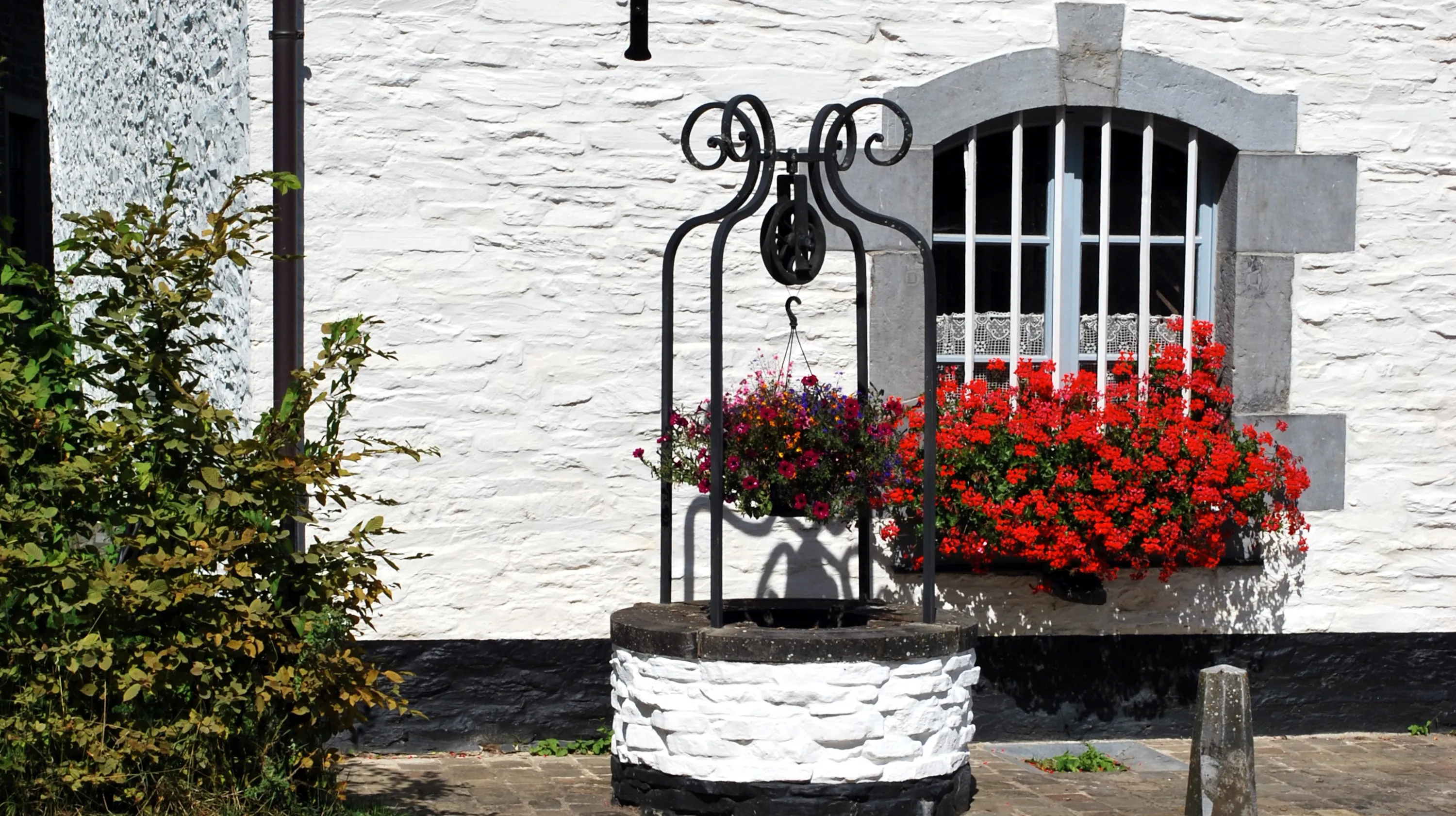 Détail de la façade de la Blanche Maison en période estivale montrant un puits à margelle en moellons chaulés, surmonté d'une ferronnerie à volutes, ainsi qu’une fenêtre à barreaux verticaux encadrée de claveaux en pierre bleue et agrémentée d'une jardinière de géraniums rouges. Ⓒ 2026 CC BY-NC 4.0 Gilles Pirlot de Corbion