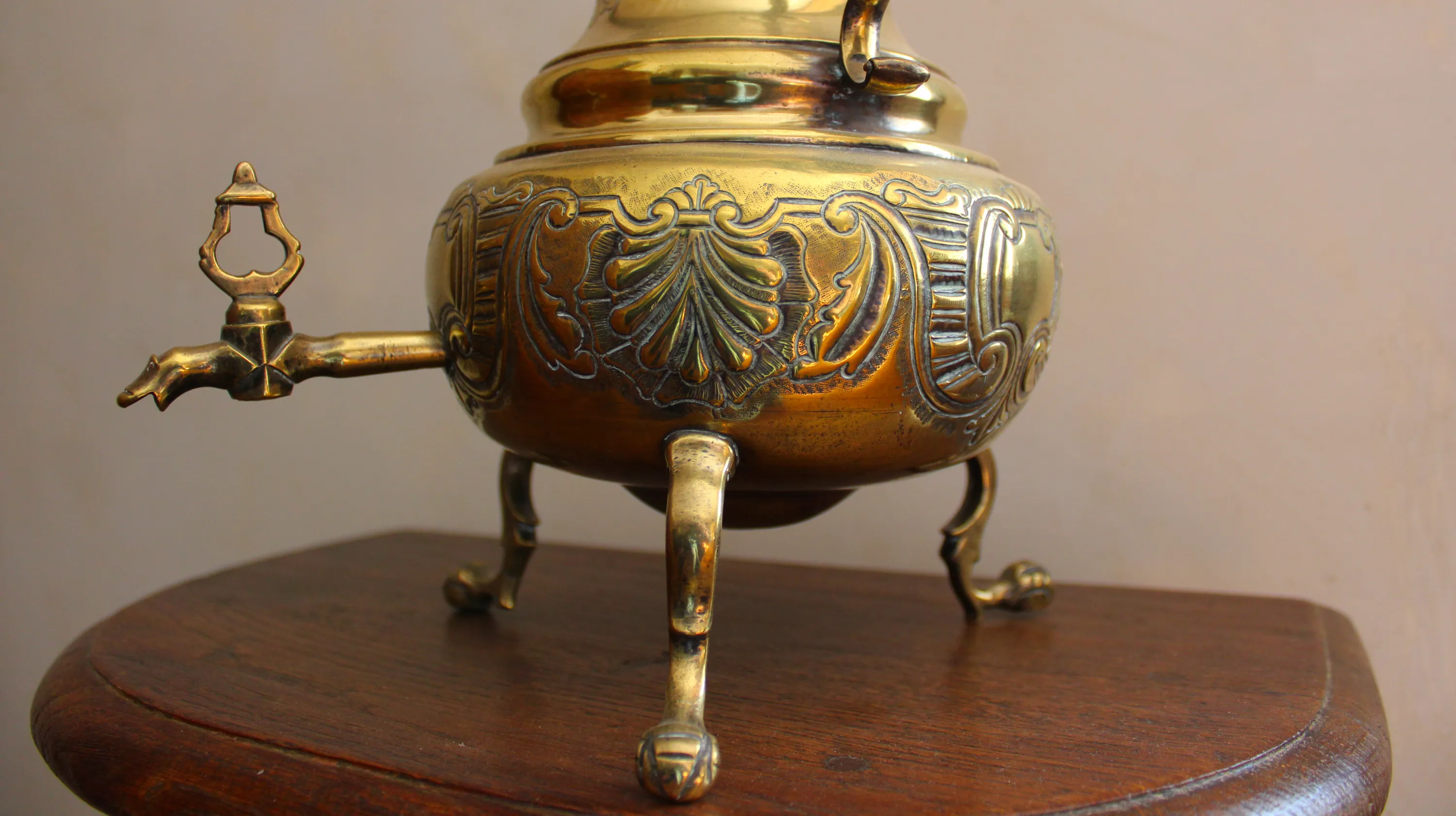 Détail d’une fontaine de table à double cambrure en laiton repoussé et gravé équipée d’un robinet dont la clef est en forme de lyre, datant des années 1730-1750 et originaire des Pays-Bas ou de Liège, présentant une panse basse ornée de coquilles encadrées de rinceaux de style Louis XIV alternant avec des cartouches godronnés et reposant sur trois pieds stylisés. Ⓒ 2026 CC BY-NC 4.0 Gilles Pirlot de Corbion