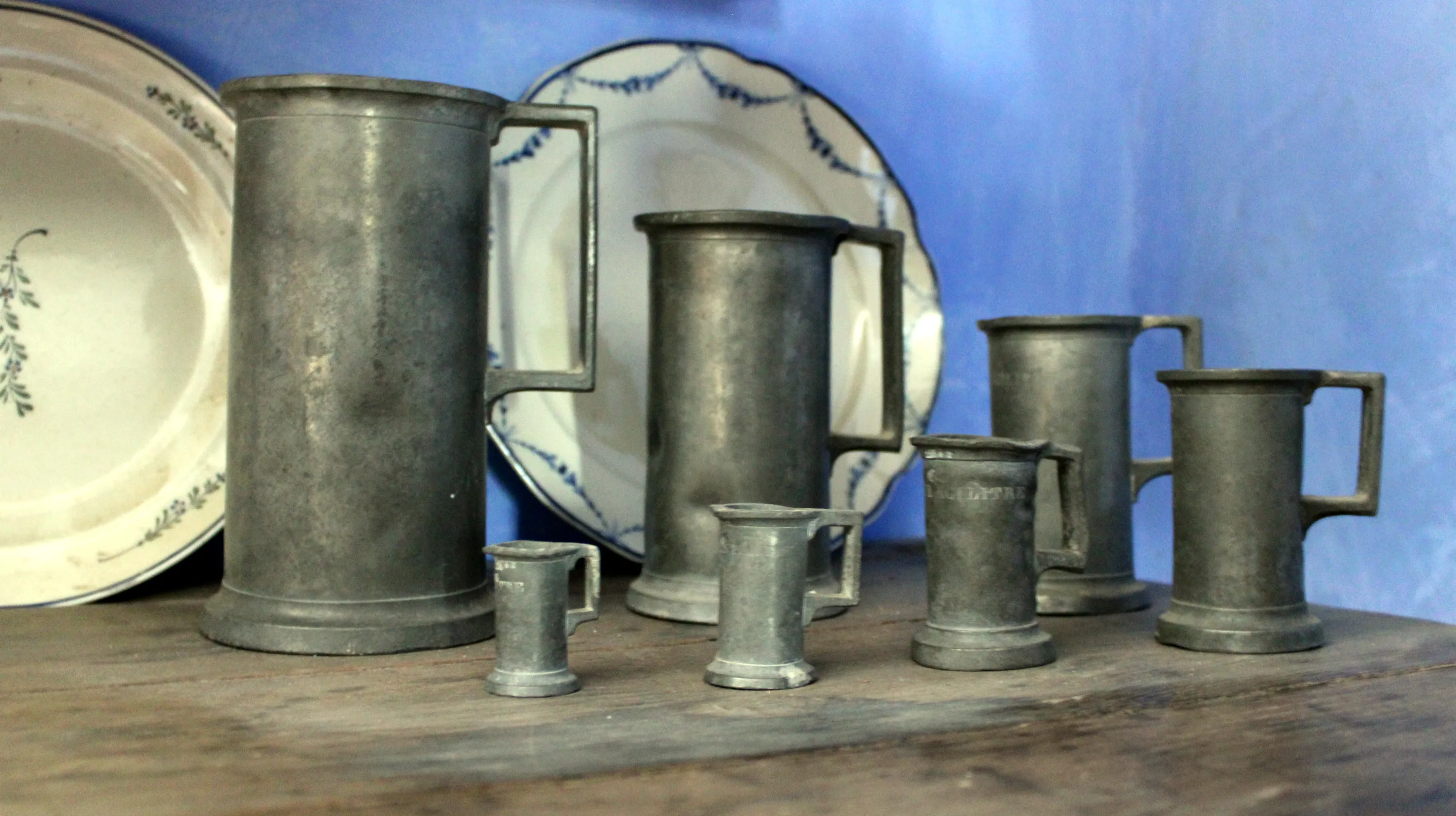 Sept récipients cylindriques en étain munis d’une anse rectangulaire fixée à mi-hauteur et d’un léger pied évasé, formant une série graduée, ainsi que deux assiettes datant de la fin du XVIIIe siècle en faïence fine de la manufacture Boch à Septfontaines, l’une décorée d’un motif végétal vert et noir et l’autre ornée de guirlandes et de festons bleus, sont posés sur un meuble de rangement en chêne foncé du XIXe siècle dans la cuisine de la Blanche Maison. Ⓒ 2026 CC BY-NC 4.0 Gilles Pirlot de Corbion