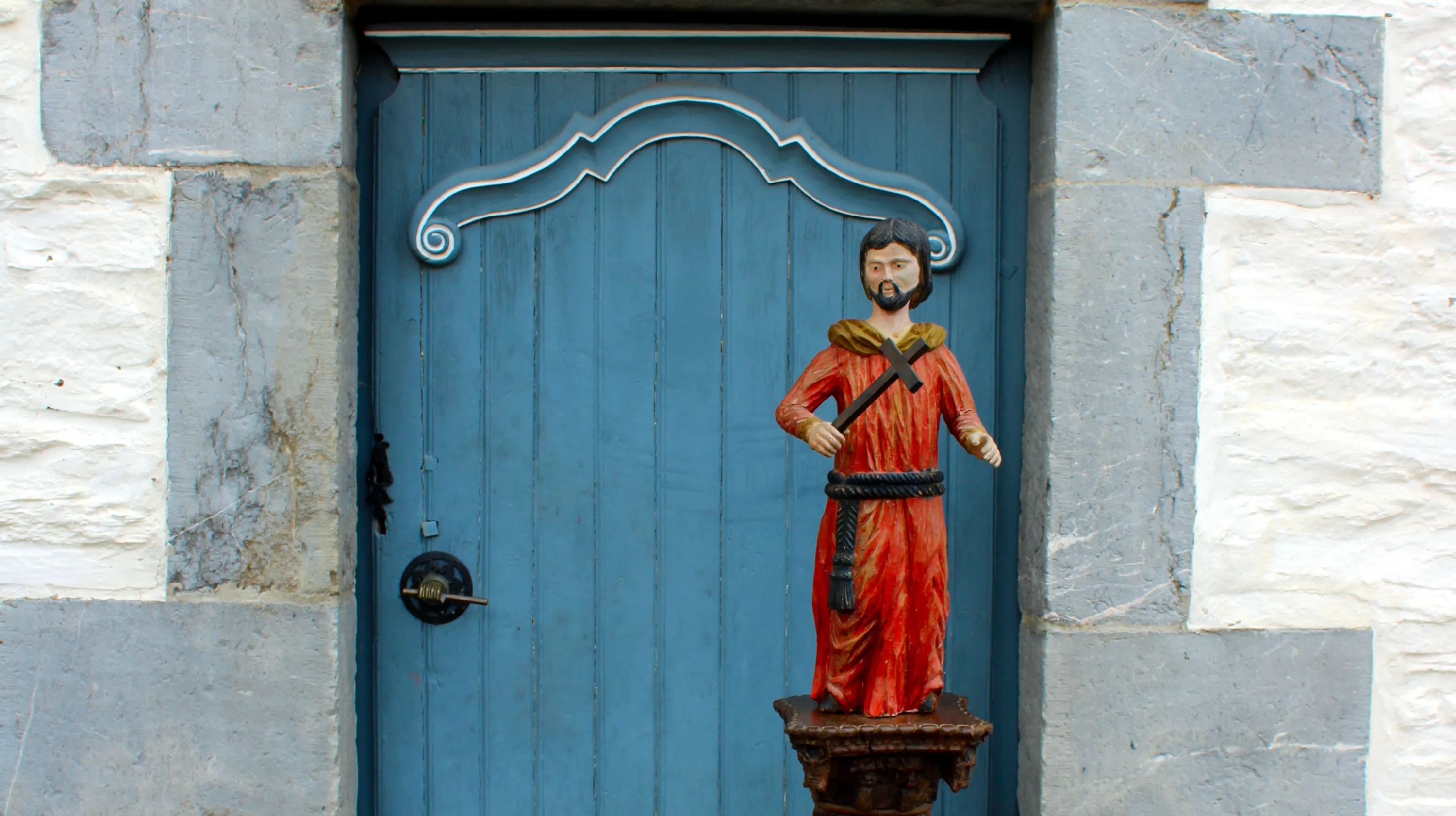 Posée sur une colonne décorée de torsades de fleurs et de fruits devant la porte d’entrée de la Blanche Maison à Nisramont, statue polychrome du XVIIIe siècle de facture naïve représentant saint François d'Assise debout, nu-tête et barbu portant un froc de moine rouge noué à la taille par une cordelière. Ⓒ 2026 CC BY-NC 4.0 Gilles Pirlot de Corbion