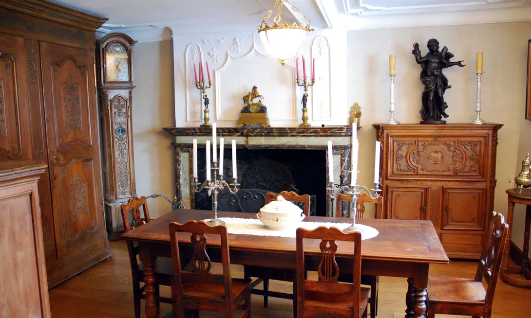 Salle d’apparat avec une cheminée, en bois peint en faux marbre, surmontée d’un trumeau en stuc orné de moulures qu’accompagnent des merles et des pampres. Les meubles consistent en un buffet, une garde robe, une gaine d’horloge et un meuble à abattant surmonté d’une grande statue de Saint Sébastien. Trois ateliers de menuiserie régionaux sont ainsi représentés. Une grande table au centre de la pièce permet d’accueillir les repas de famille. Ⓒ 2024 CC BY-NC 4.0 Gilles Pirlot de Corbion