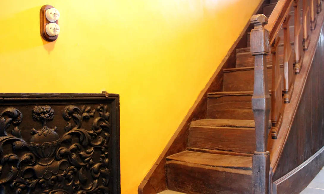 Du couloir peint en jaune, superposé à celui de la cave, démarre l’escalier en chêne menant à l’étage. Sa rampe à balustres et son départ d’escalier, un peu maigre mais typique de la région, sont de style Louis XIV. La taque de cheminée à décor héraldique en fort relief arbore les armes d’alliance Piret de Sainte-Ode et Poschet surmontées d’un heaume avec cimier et de lambrequins. Ⓒ 2024 CC BY-NC 4.0 Gilles Pirlot de Corbion