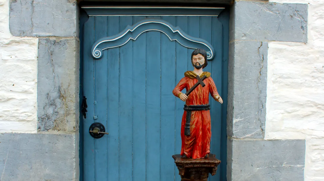 Posée sur une colonne décorée de torsades de fleurs et de fruits devant la porte d’entrée de la Blanche Maison à Nisramont, statue polychrome du XVIIIe siècle de facture naïve représentant saint François d'Assise debout, nu-tête et barbu portant un froc de moine rouge noué à la taille par une cordelière. Ⓒ 2026 CC BY-NC 4.0 Gilles Pirlot de Corbion
